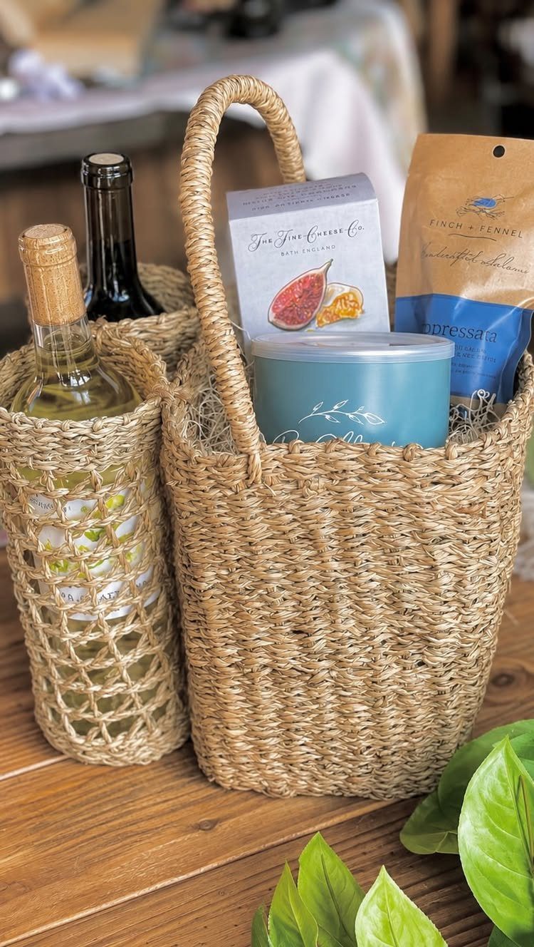 Woven straw gift basket with wine bottles, a blue candle, and gourmet snacks.