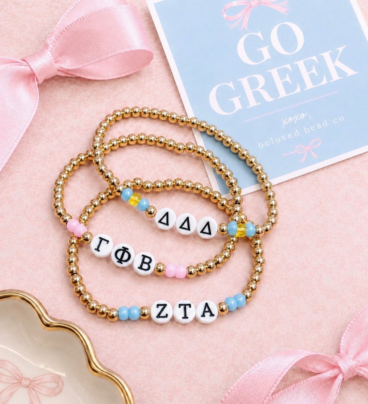 Three gold beaded bracelets with Greek letters on a pink background with ribbons.