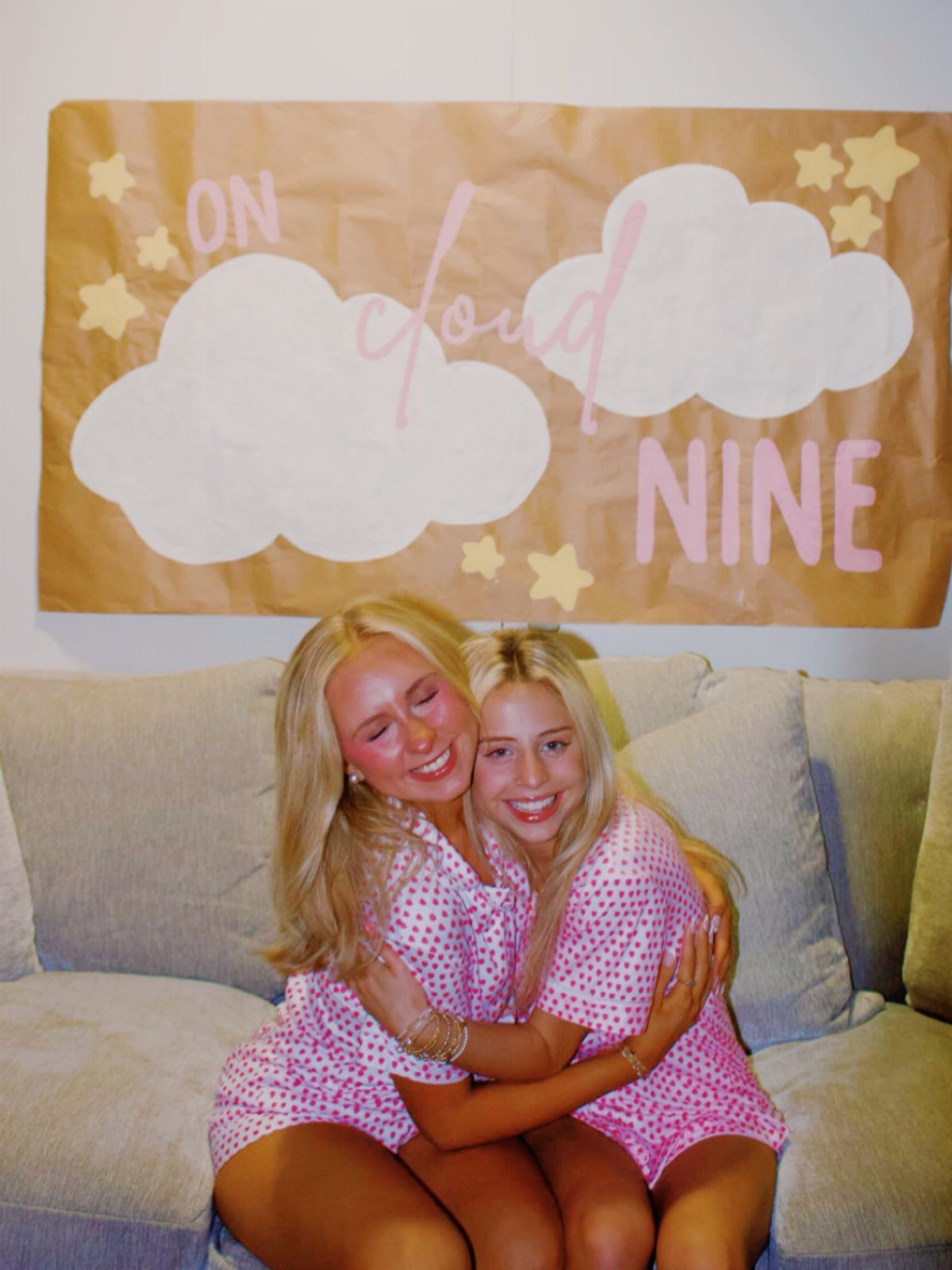 Two women in matching pink polka dot pajamas hugging on a grey sofa.