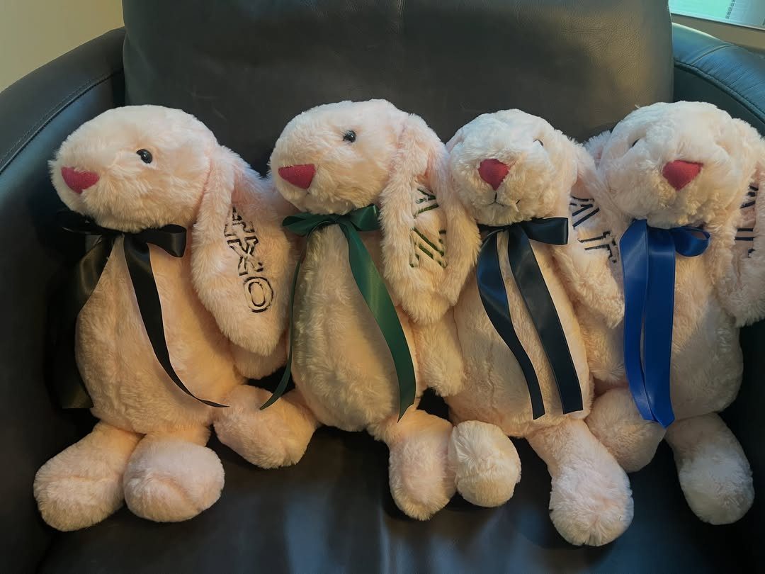 Four plush pink bunnies with colorful ribbons sitting on a grey leather chair.