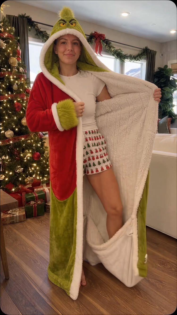 Woman wearing a Grinch-themed hooded robe standing next to a decorated Christmas tree.