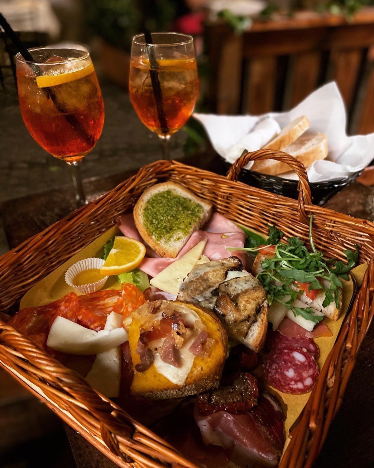 Wicker basket filled with Italian meats and cheeses next to two Aperol Spritz cocktails.