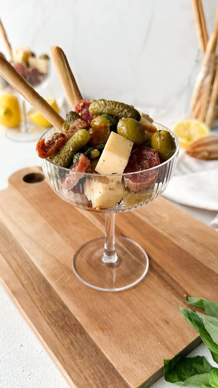 Glass bowl of olives, pickles, and cheese on a wooden cutting board.