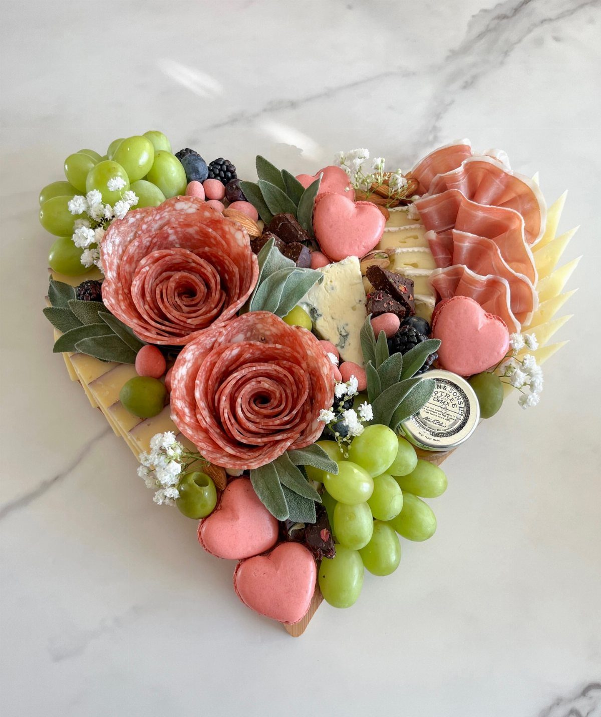 Heart-shaped charcuterie board with salami roses, green grapes, and pink heart chocolates.