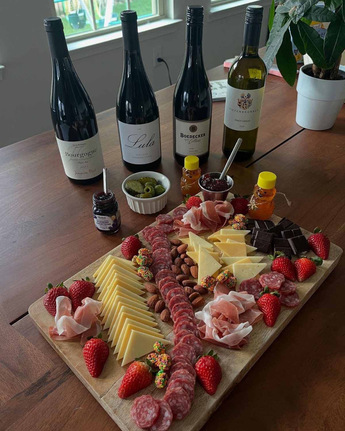 Four wine bottles standing behind a wooden board with meats, cheeses, and strawberries.