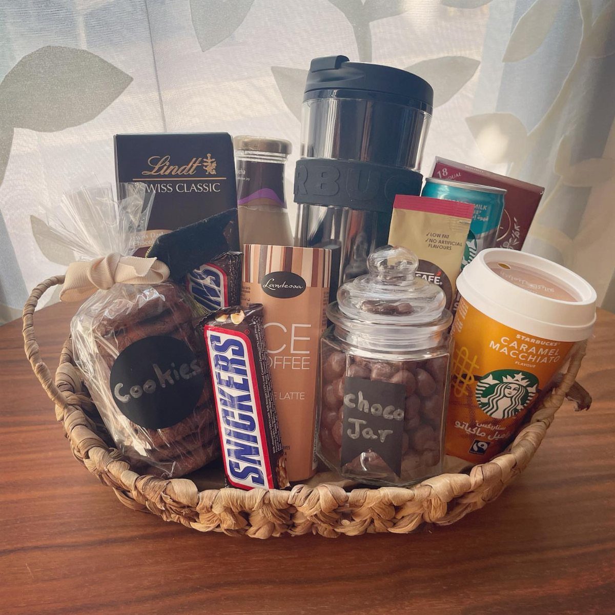 Christmas Gift Baskets for Family: 7 Ideas for Every Age 4 Woven gift basket containing coffee drinks, chocolates, and snacks on a wooden surface.
