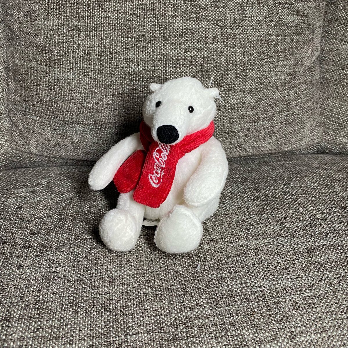 Coca-Cola Christmas Gift Ideas: 7 Picks for Fans 2 White plush polar bear wearing a red Coca-Cola scarf on grey fabric.