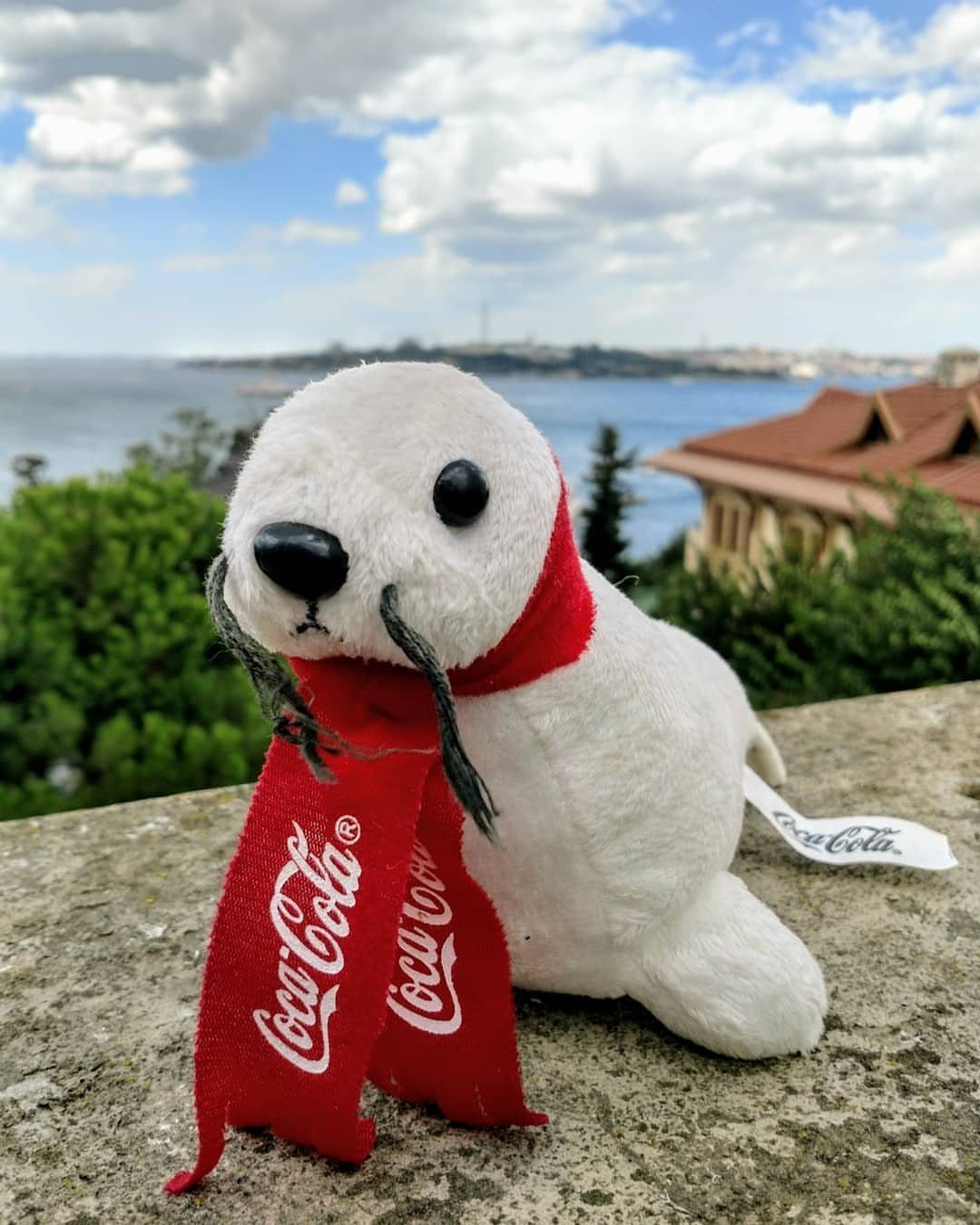Coca-Cola Christmas Gift Ideas: 7 Picks for Fans 4 White plush seal wearing a red Coca-Cola scarf on a stone ledge outdoors.