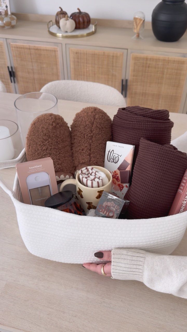 10 Cozy Gift Basket Ideas, from Spa Sets to Pumpkin Spice 9 White woven gift basket containing brown slippers, a blanket, and chocolate treats.