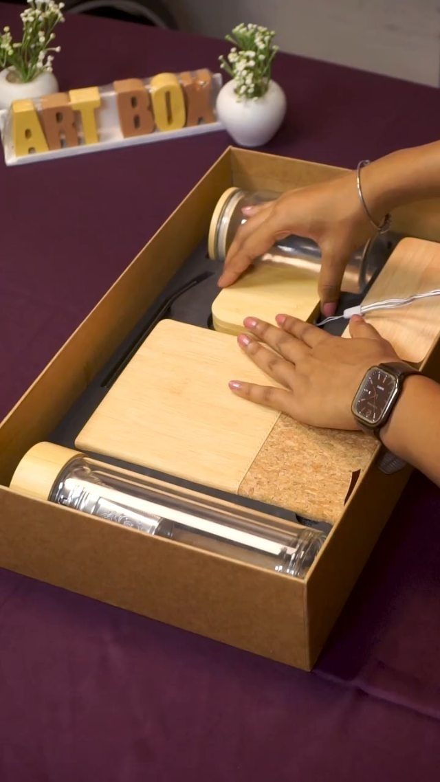 Hands arranging glass water bottles and wooden accessories in a brown gift box.