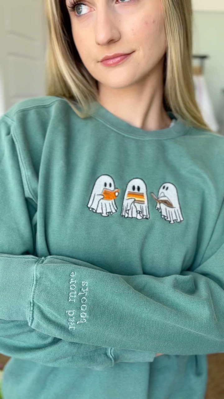 13 Fall Gifts for Women, From Cozy Socks to Cookbooks 2 Woman wearing a sage green sweatshirt with ghost graphics and book-themed sleeve text.
