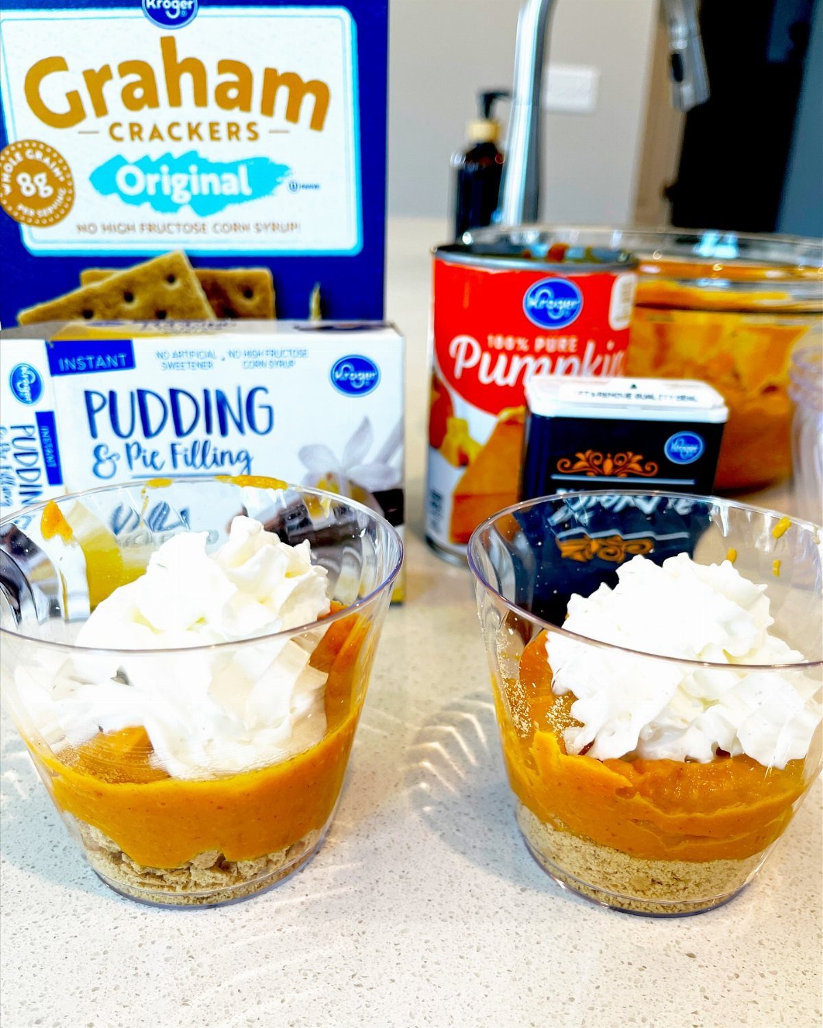 Layered pumpkin desserts in plastic cups with ingredients in the background.