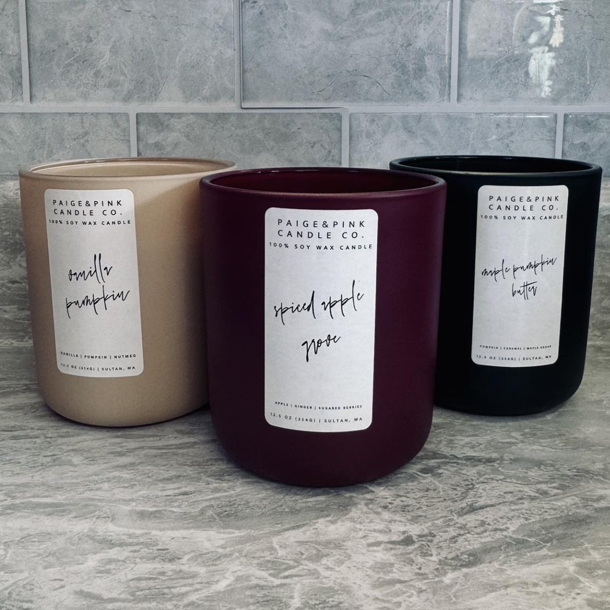 Three scented soy wax candles in tan, burgundy, and black jars on marble.