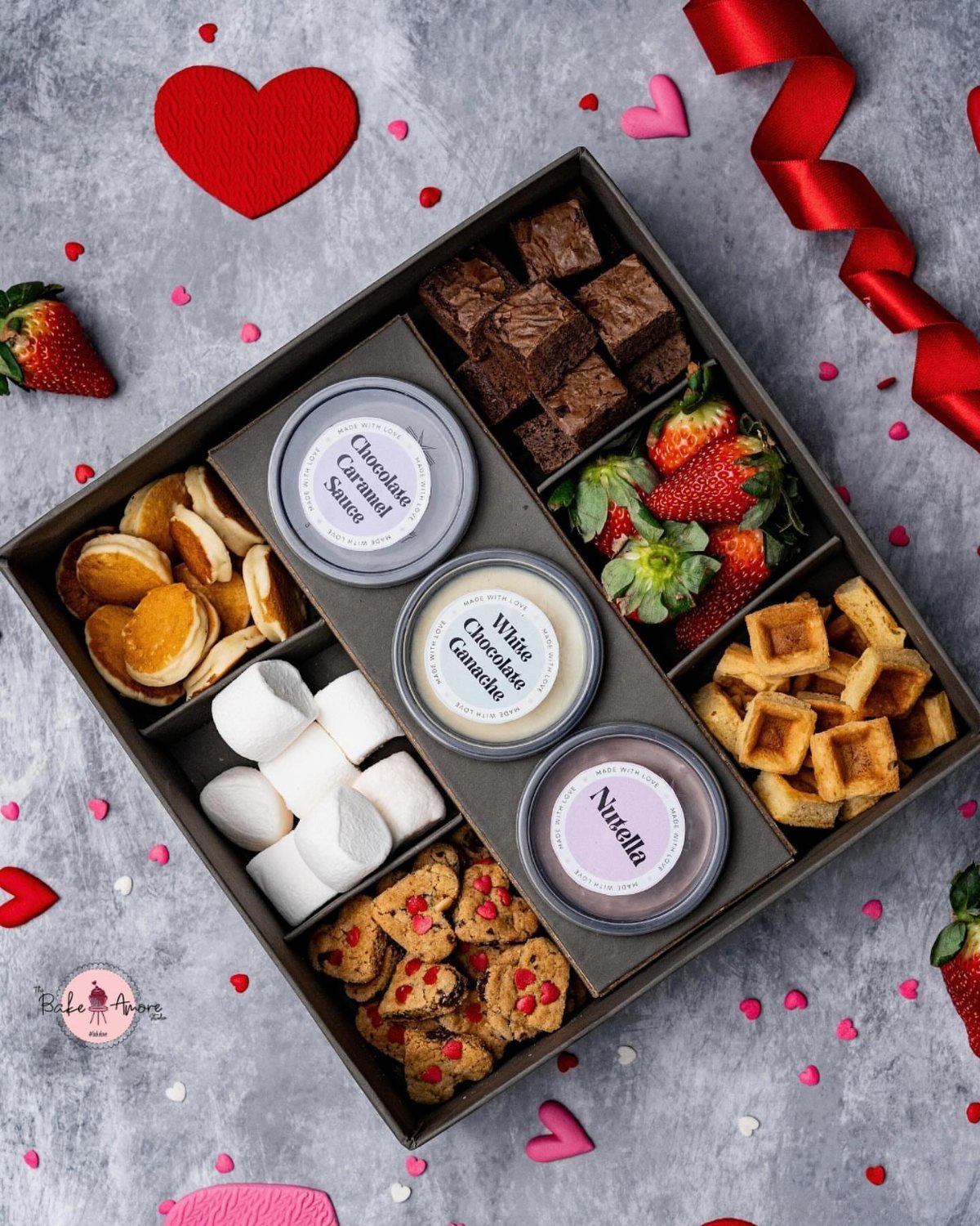 Dessert gift box with brownies, strawberries, waffles, and dipping sauces surrounded by red hearts.