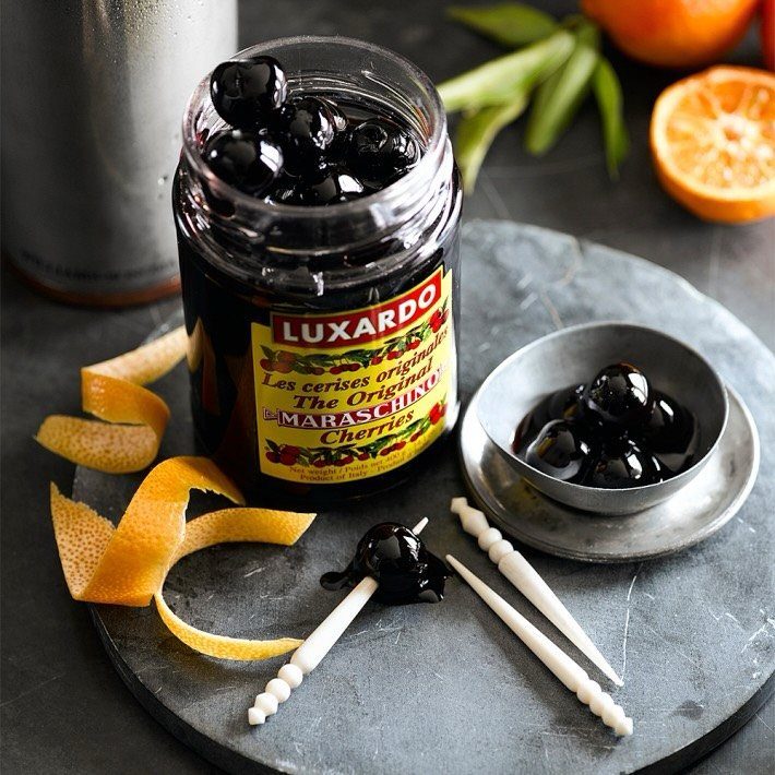 Luxardo Maraschino cherries in a jar and bowl with orange peels on stone.