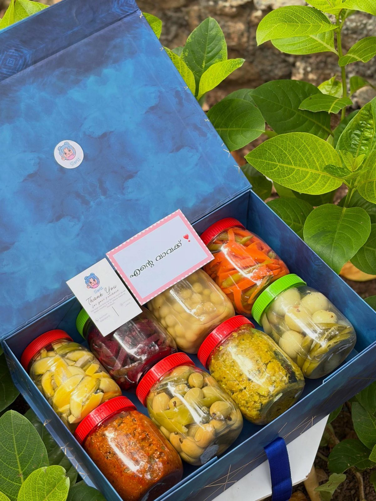Blue gift box filled with assorted jars of colorful pickled vegetables and preserves.