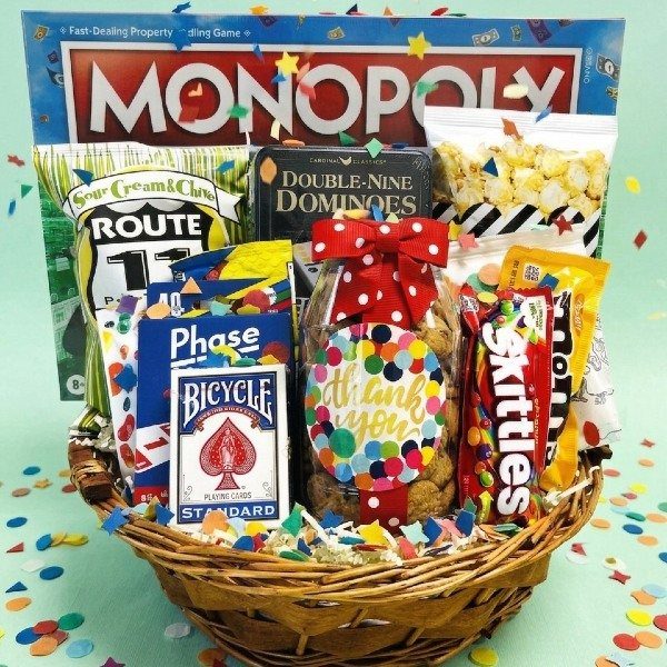 Wicker gift basket containing Monopoly, dominoes, playing cards, and various snacks with confetti.