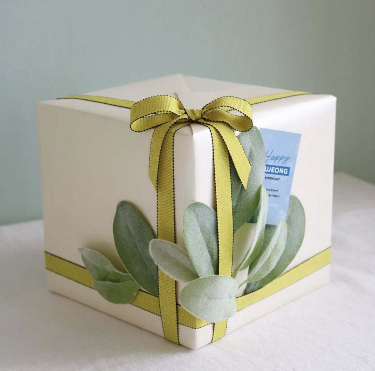 Cream gift box with lime green ribbon bow and silvery-green foliage leaves.