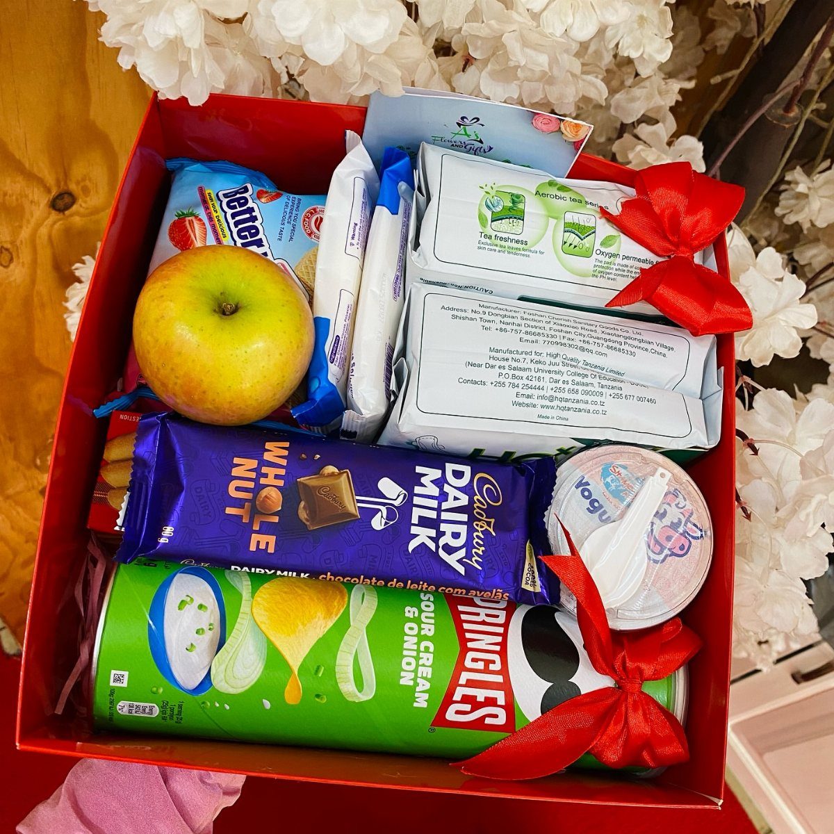 Red gift box filled with snacks, chocolate, and an apple with red ribbons.