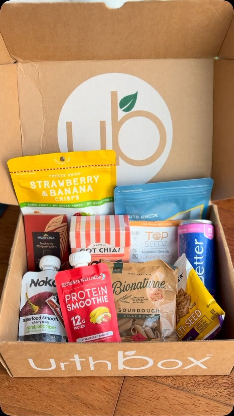 Open cardboard subscription box filled with various healthy packaged snacks and smoothies.