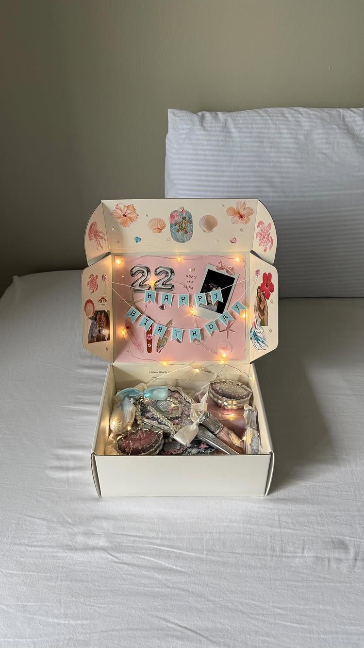 Open white gift box with personalized birthday banner, fairy lights, and assorted treats.