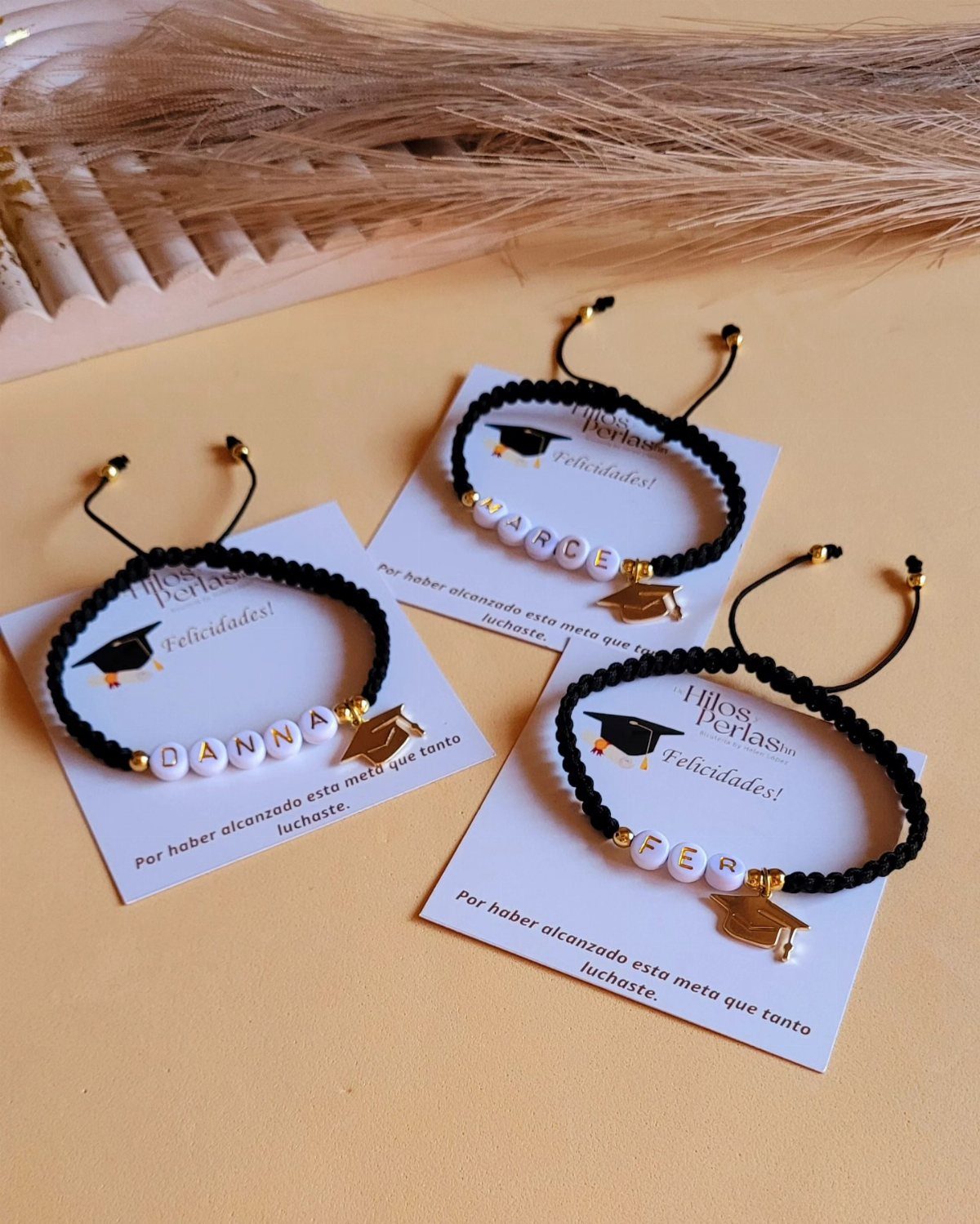 Three black braided friendship bracelets with name beads and gold graduation cap charms.
