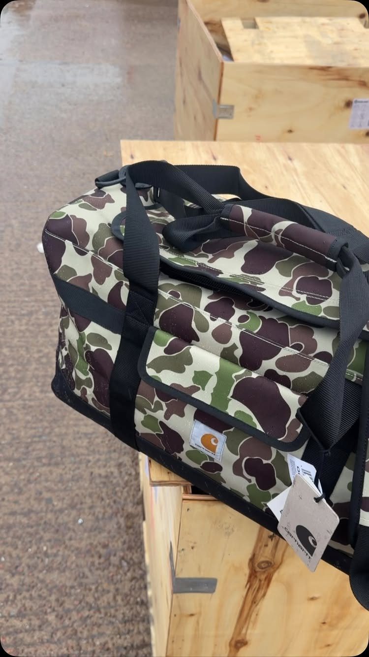 Camouflage Carhartt duffel bag with black straps sitting on a wooden crate.