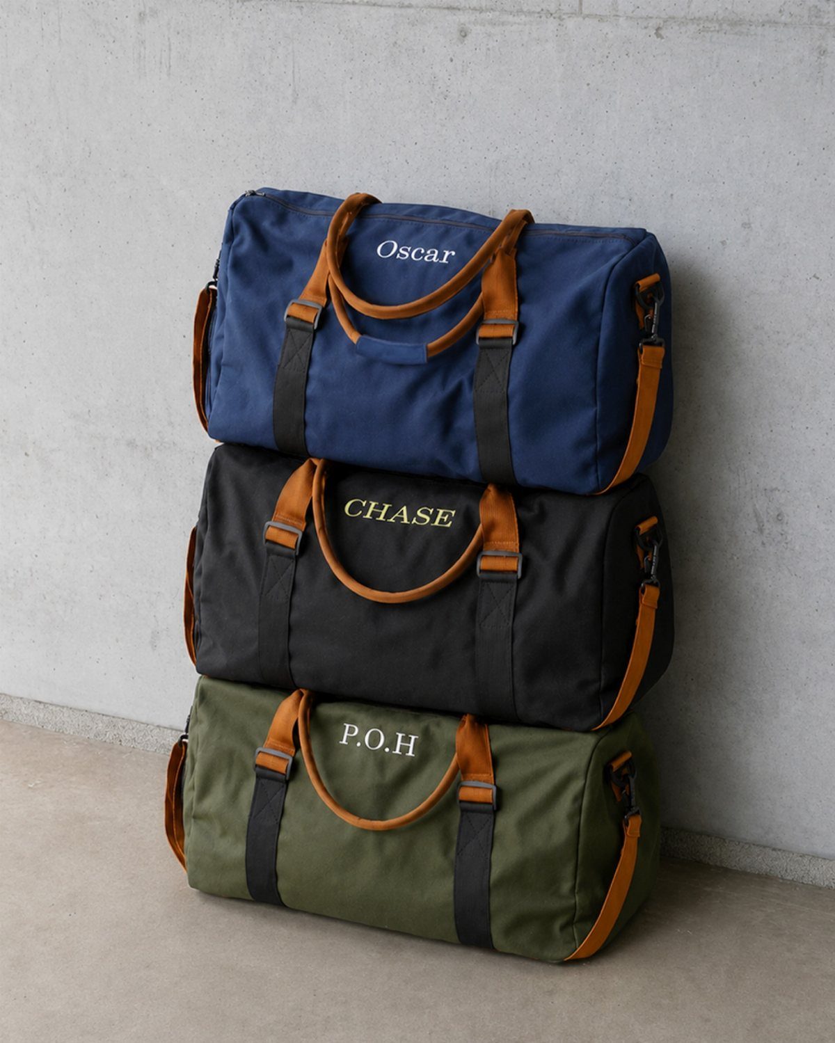 Three personalized canvas duffel bags in blue, black, and olive green stacked vertically.
