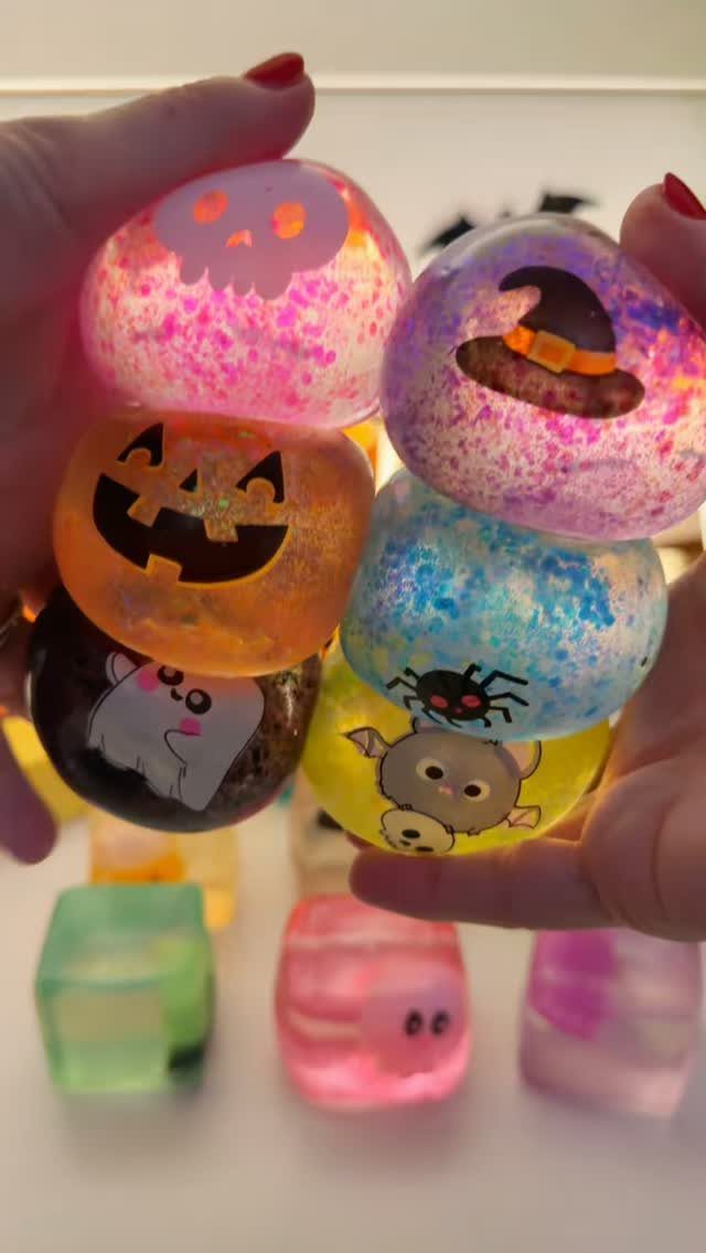 Colorful glittery Halloween stress balls with pumpkin and ghost designs held in hands.