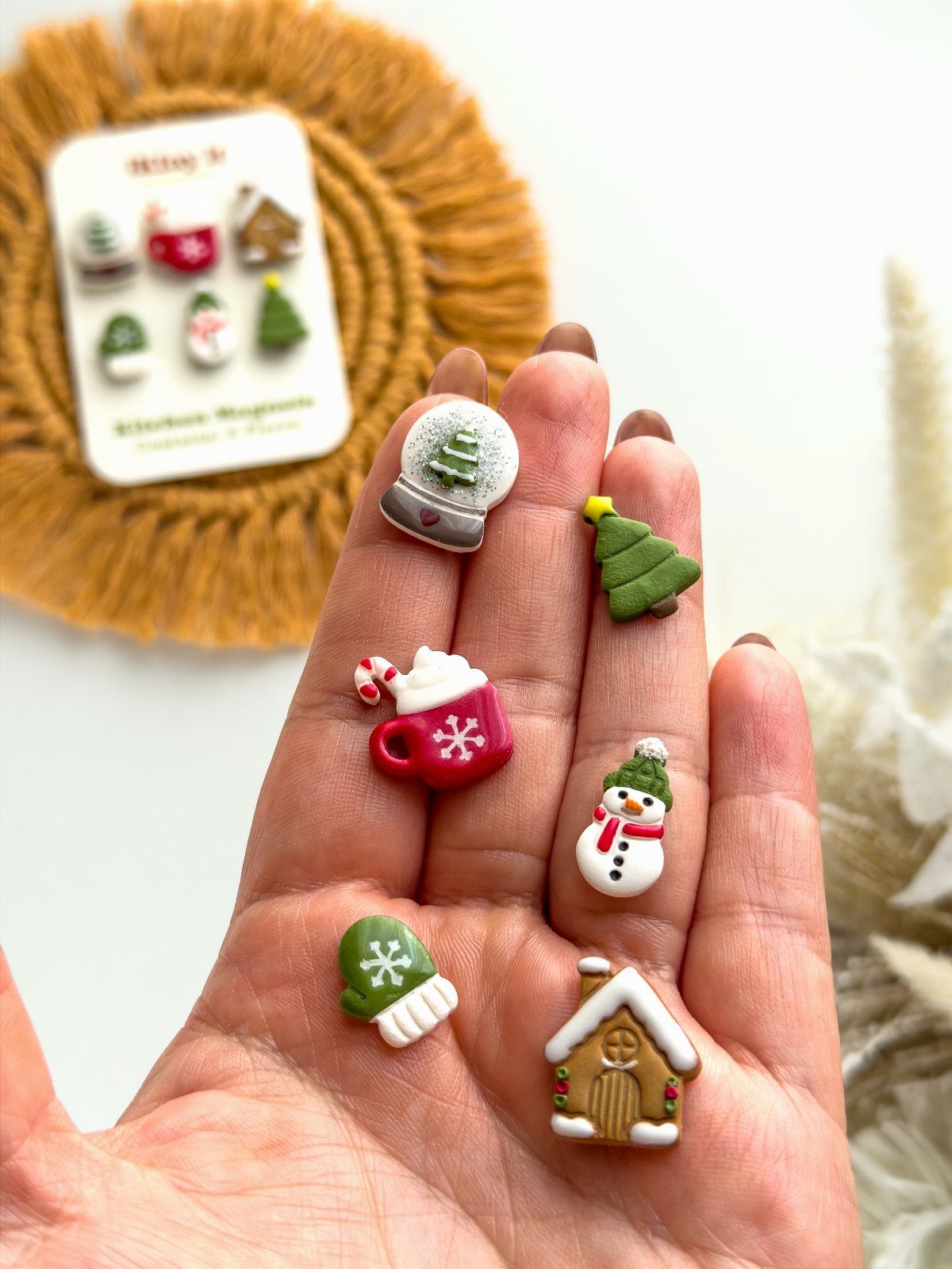 Small colorful clay Christmas magnets held in a hand, including a snowman and house.