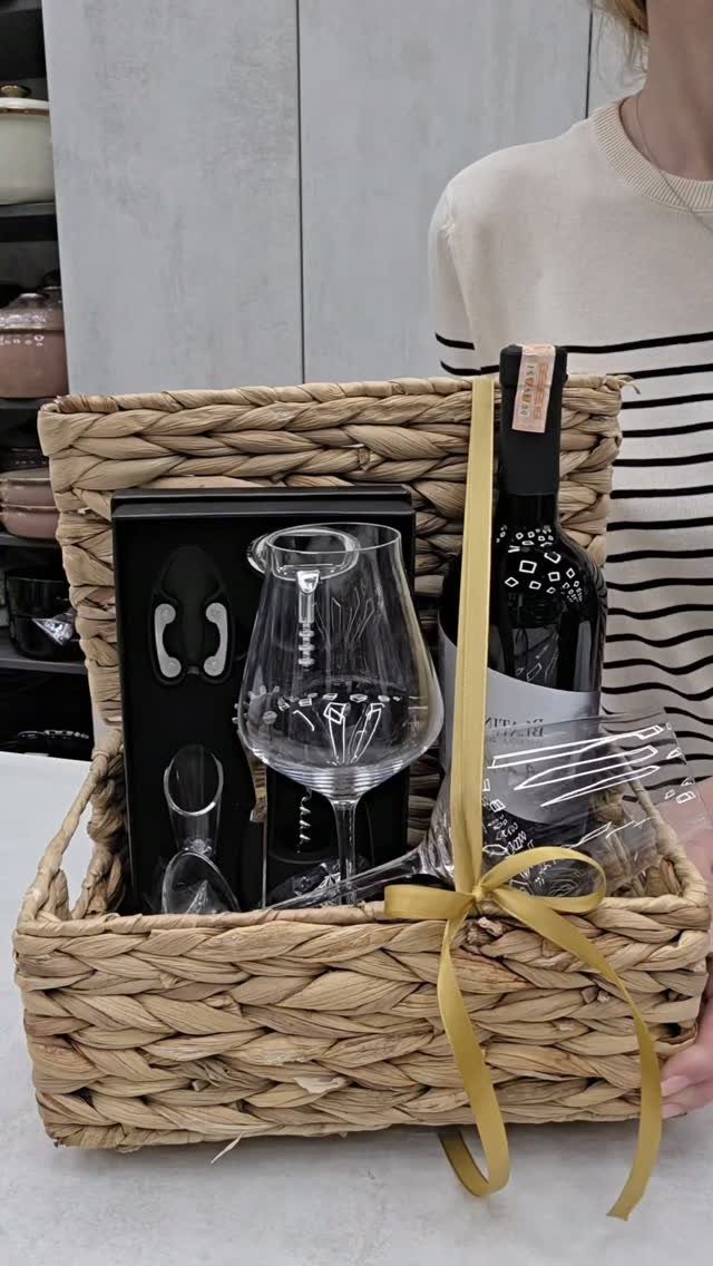 Woven wicker gift basket with red wine, wine glass, and accessories tied with gold ribbon