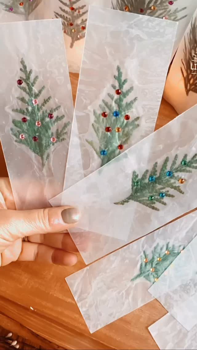 Translucent bookmarks with painted evergreen trees and colorful rhinestones held by a hand.