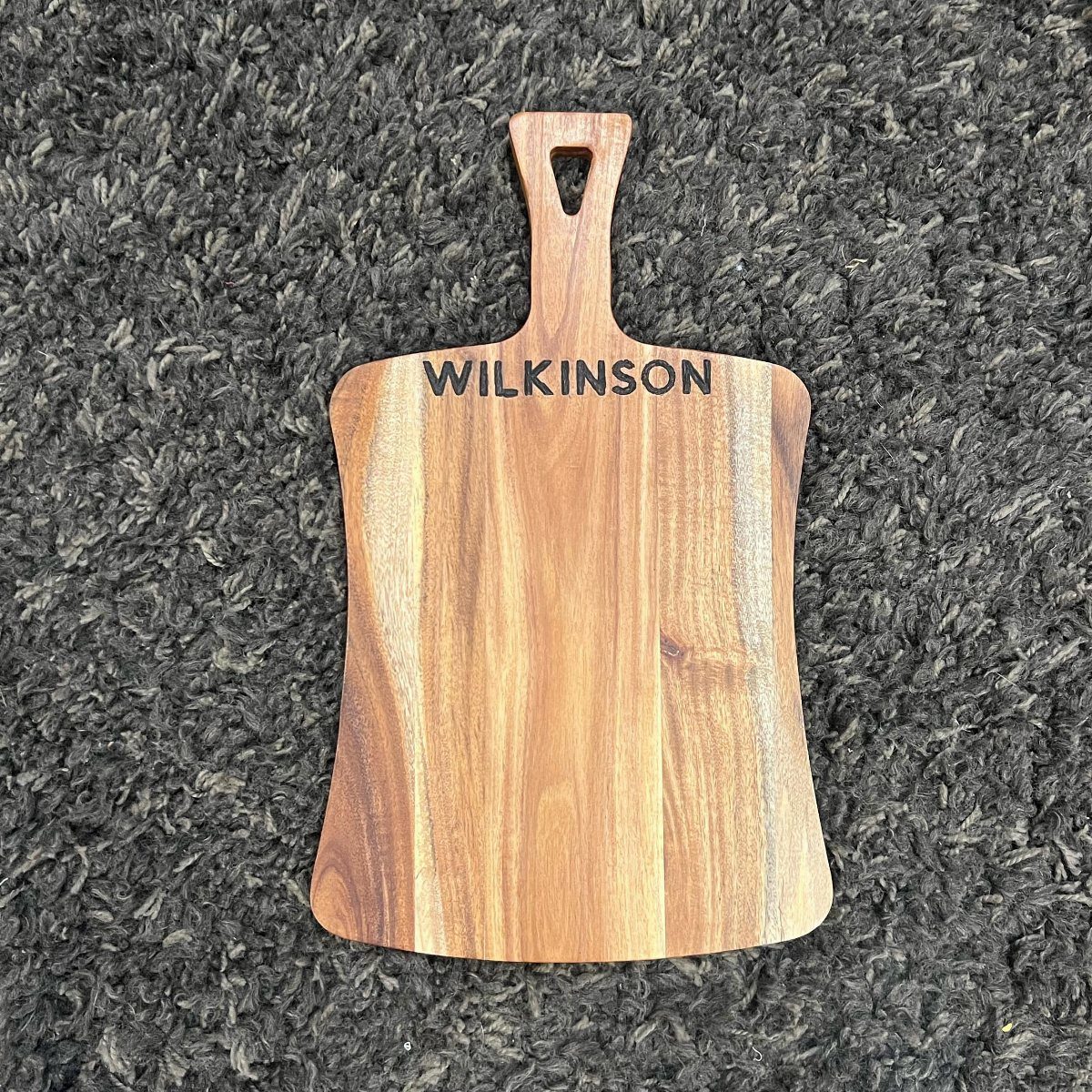 Wooden cutting board with the name WILKINSON engraved on a grey shag carpet