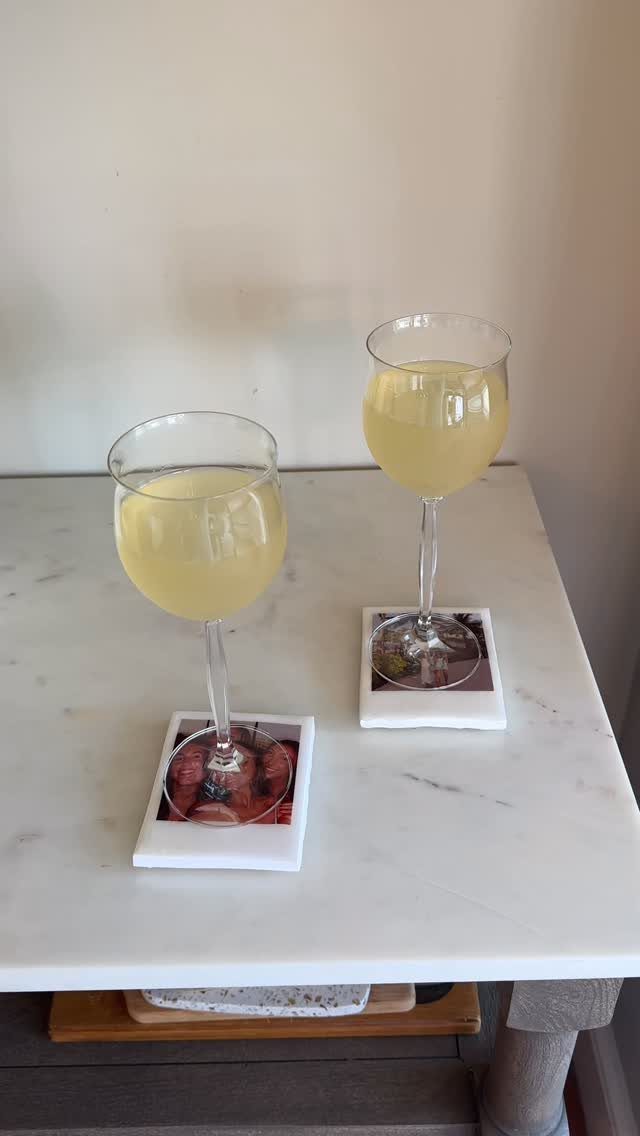 Two glasses of yellow drink on square white photo coasters on marble table