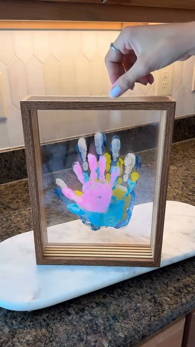 Colorful child's handprint art in a square wooden frame held by a hand.