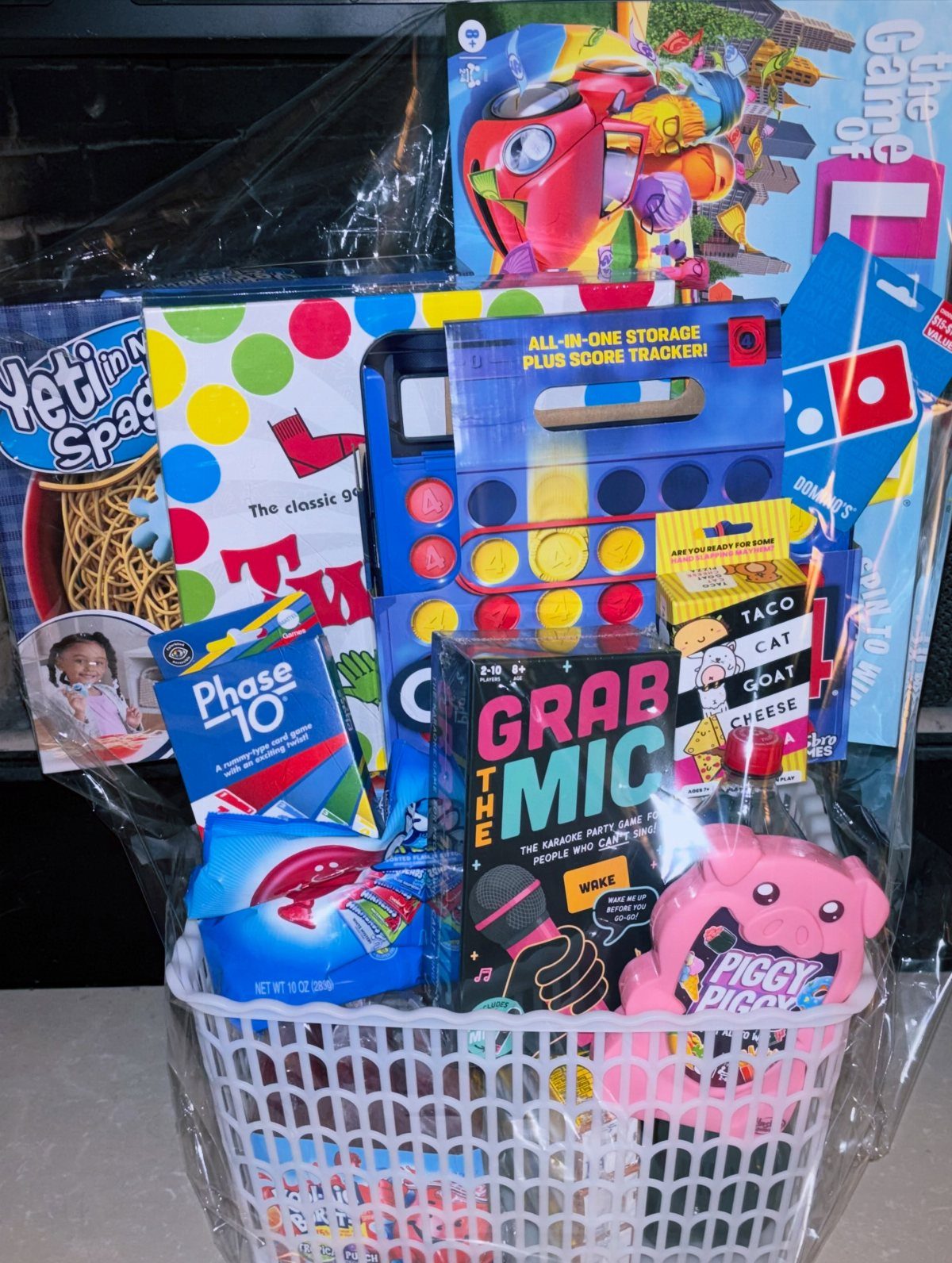 14 Household Gift Basket Ideas for Every Occasion 2 White gift basket filled with board games and snacks wrapped in clear cellophane.