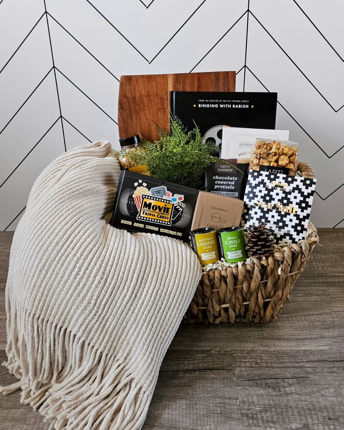 14 Household Gift Basket Ideas for Every Occasion 1 Woven gift basket with popcorn, movie snacks, a book, and a cream throw blanket.