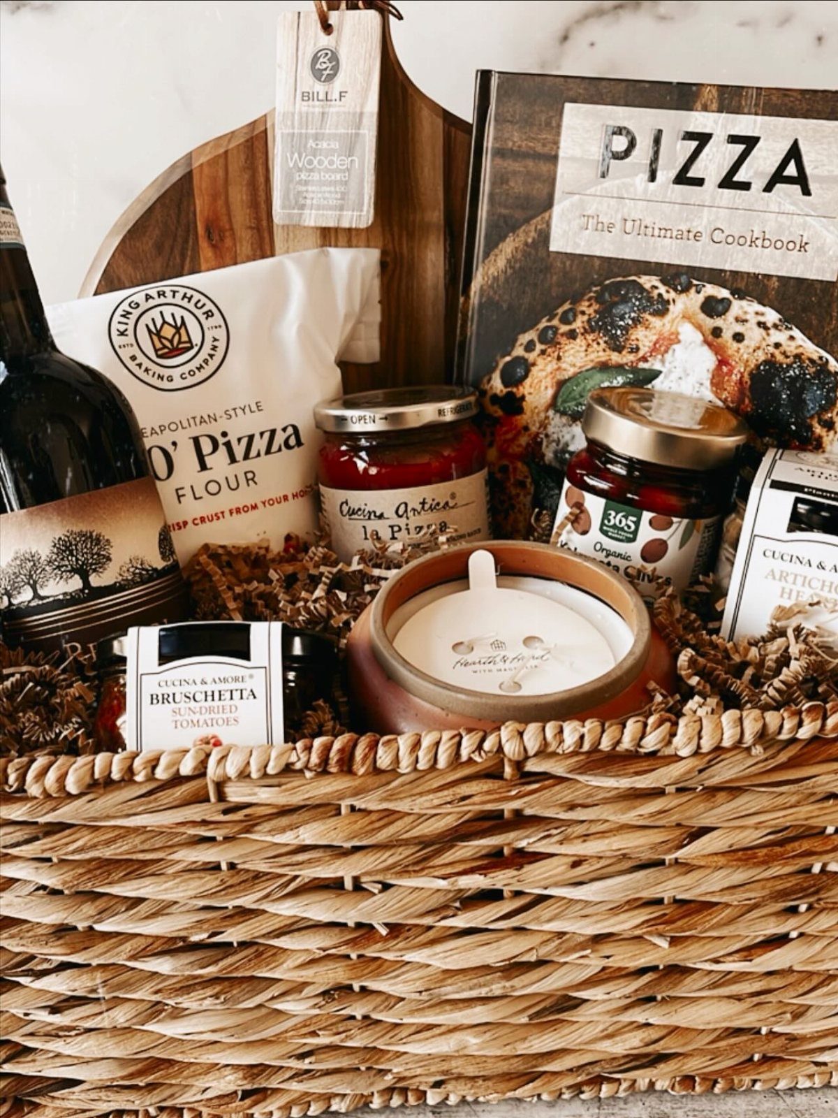 14 Household Gift Basket Ideas for Every Occasion 7 Woven gift basket containing pizza cookbook, flour, sauce, wine, and wooden cutting board.