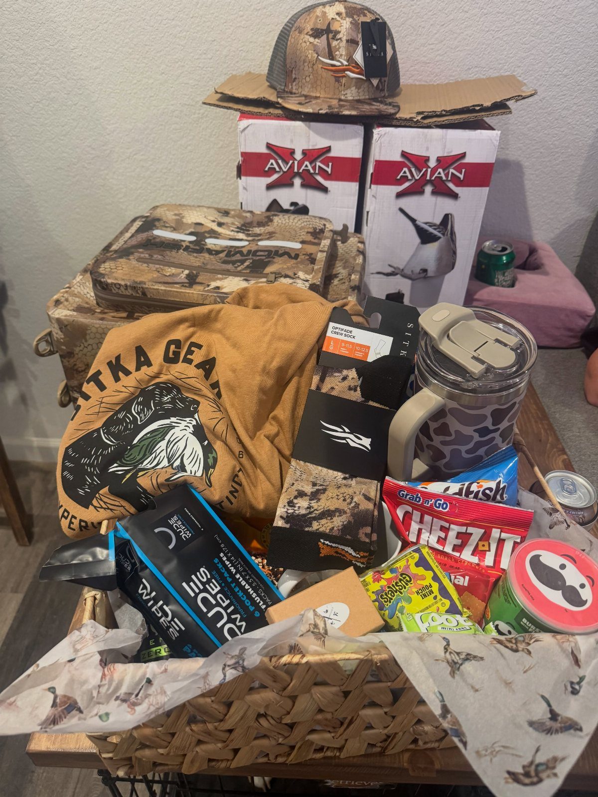 Hunting Gift Basket Ideas: 7 Options to Give This Year 4 Wicker gift basket filled with camouflage hunting gear, snacks, and a tan t-shirt.