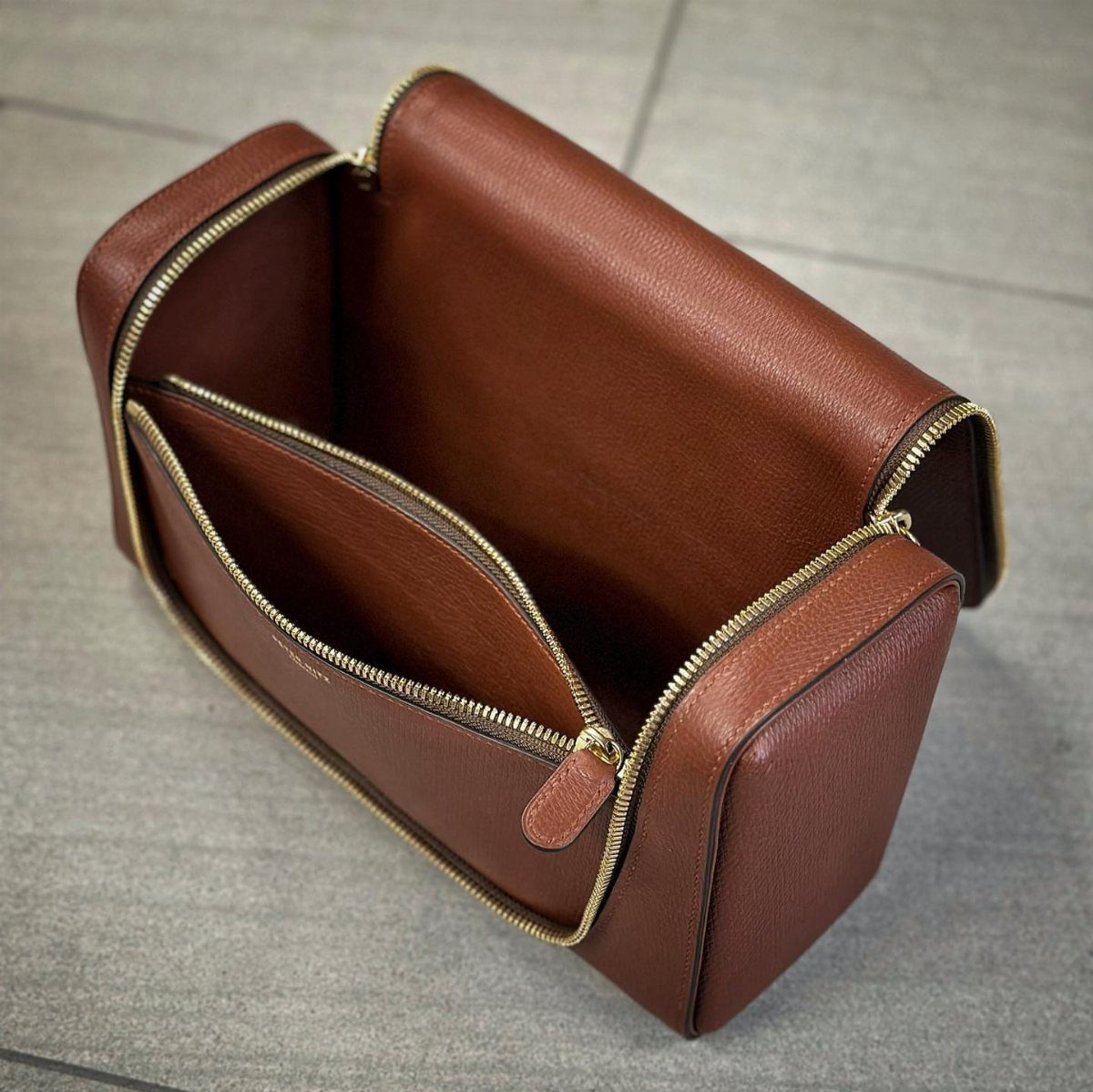 Brown leather toiletry bag with gold zippers open on a grey tile floor