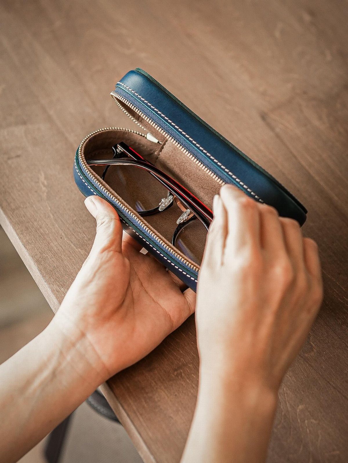 Hands holding an open blue leather glasses case on a wooden table
