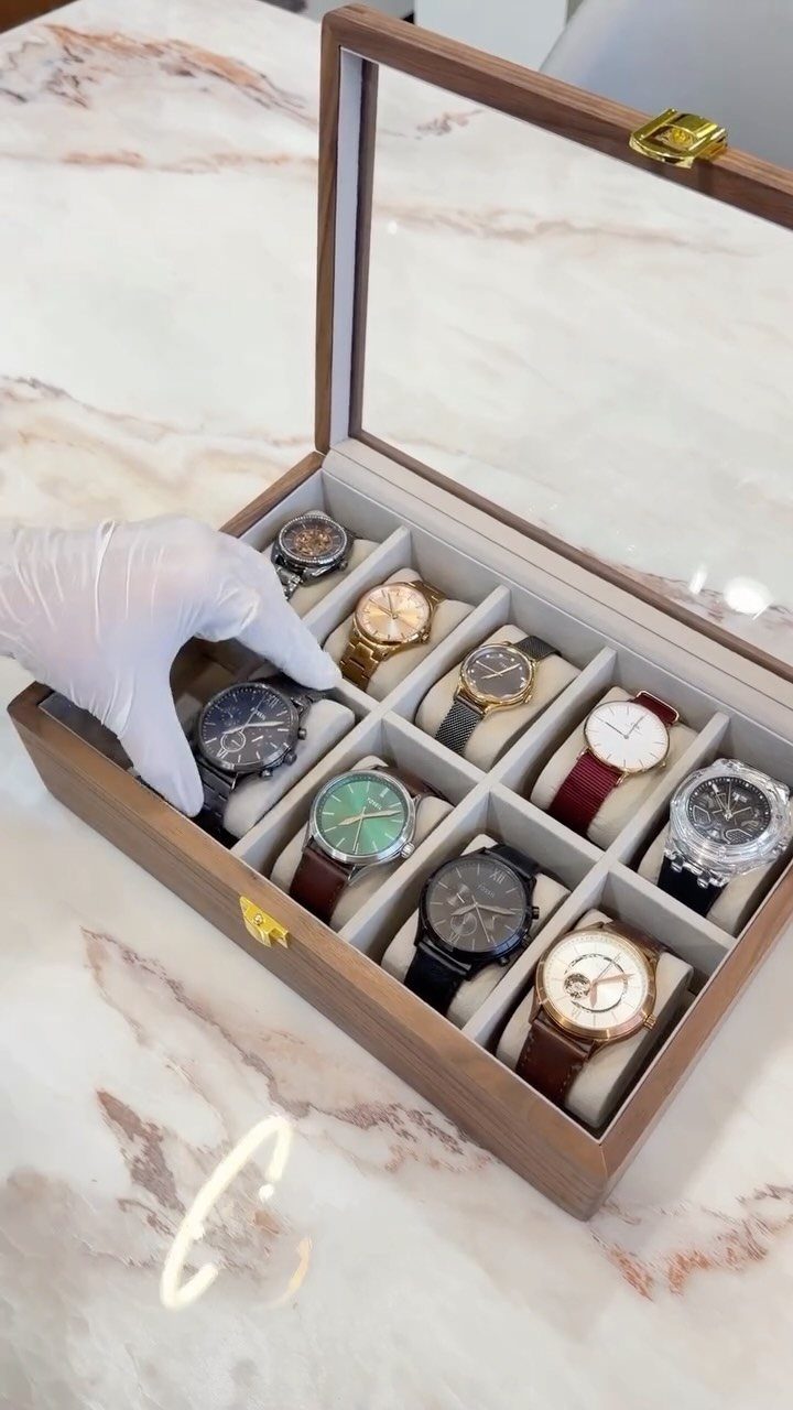 Wooden watch organizer case with glass lid holding ten luxury wristwatches on marble.
