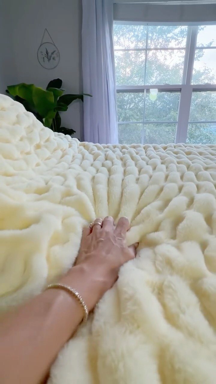 Hand resting on a thick, cream-colored chunky knit faux-fur cozy blanket on a bed.