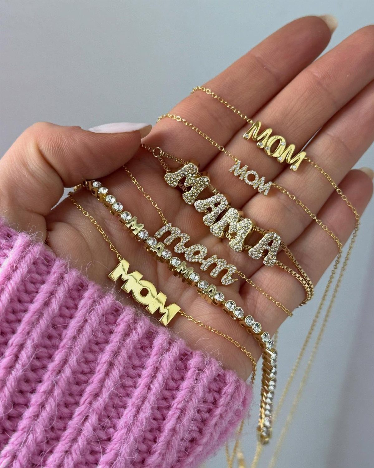 Gold necklaces with MOM and MAMA pendants held in a hand with pink sweater