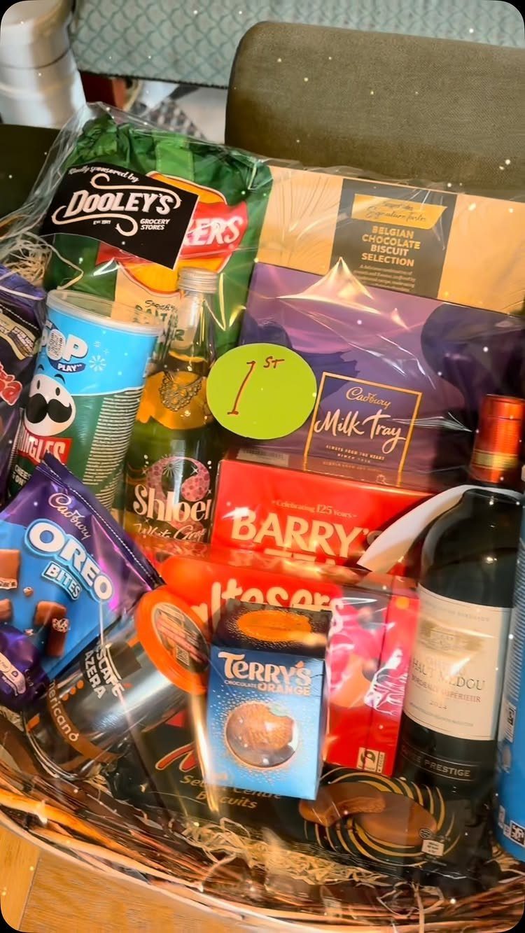 Wicker gift basket filled with assorted chocolates, wine, and snacks wrapped in cellophane.