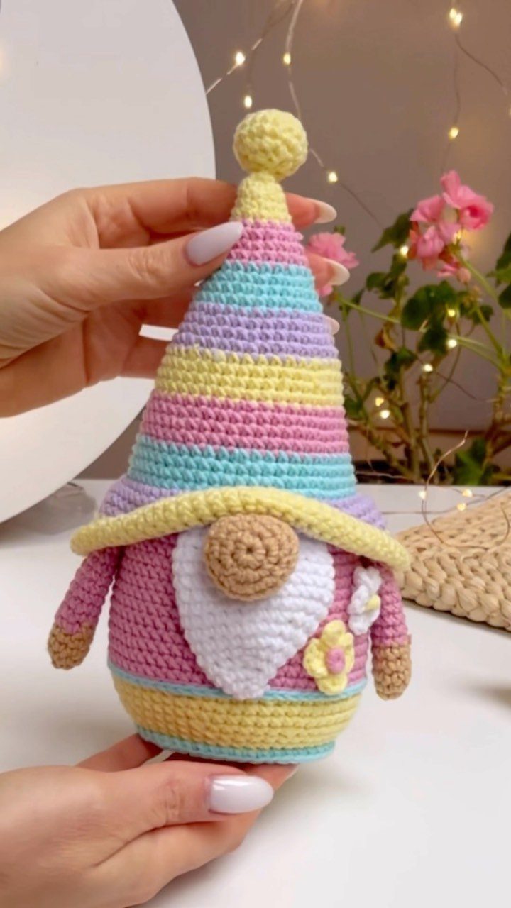 Crocheted gnome plush doll with a striped hat held by two hands.