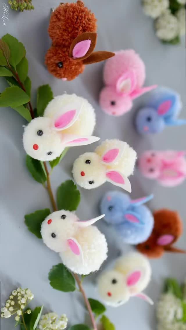 Colorful pom-pom bunnies with felt ears scattered among green leaves and white flowers.