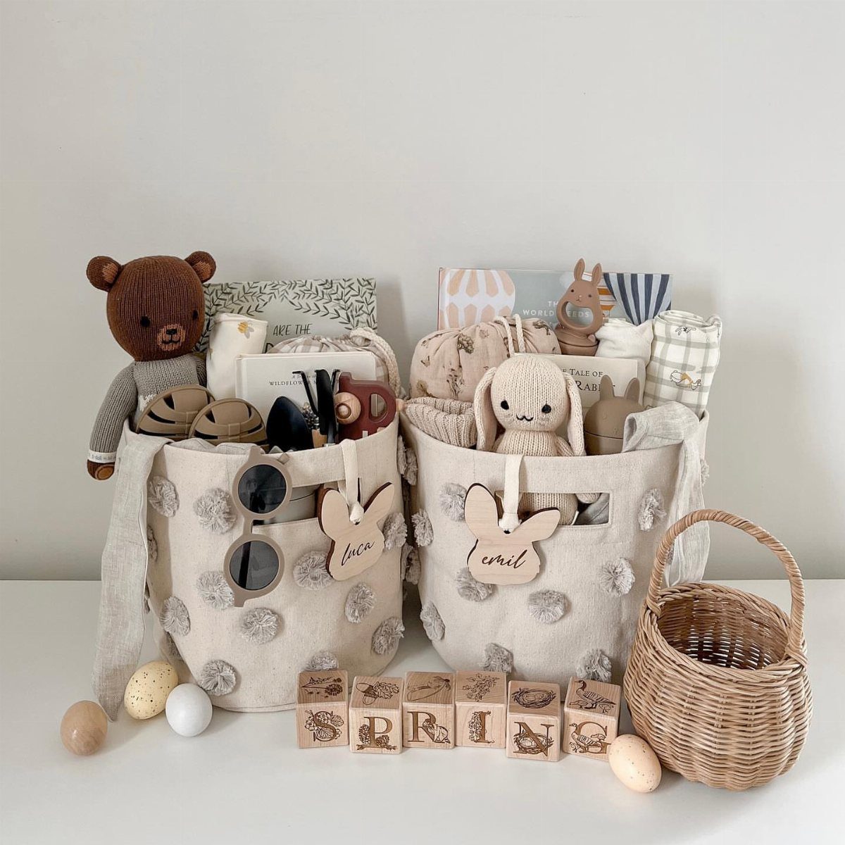Two cream fabric baskets with grey pom-poms filled with baby gifts and plush toys.