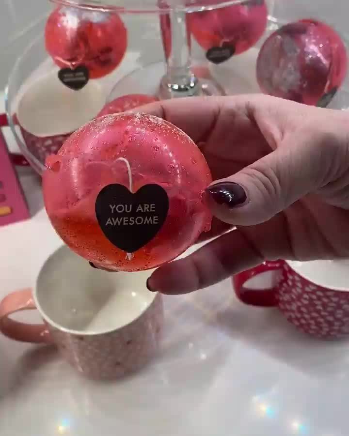 Tea Bag Gift Ideas to Make or Buy: 11 Sweet Picks 6 Red spherical tea bombs with heart labels held over white ceramic mugs.
