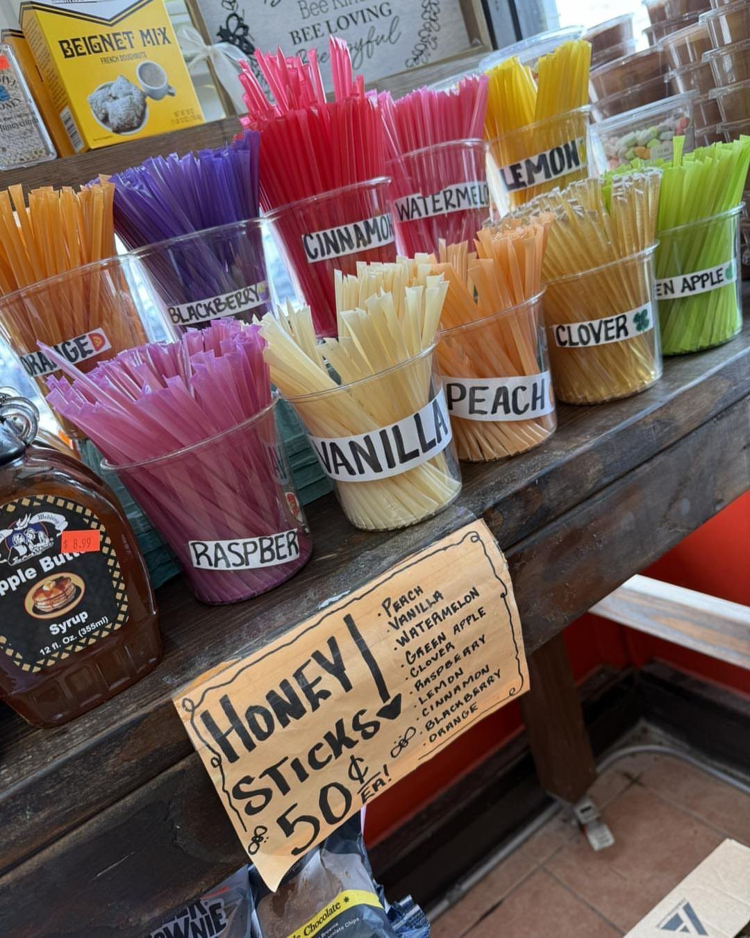 Tea Bag Gift Ideas to Make or Buy: 11 Sweet Picks 4 Colorful honey sticks in clear cups on a wooden shelf with a price sign.
